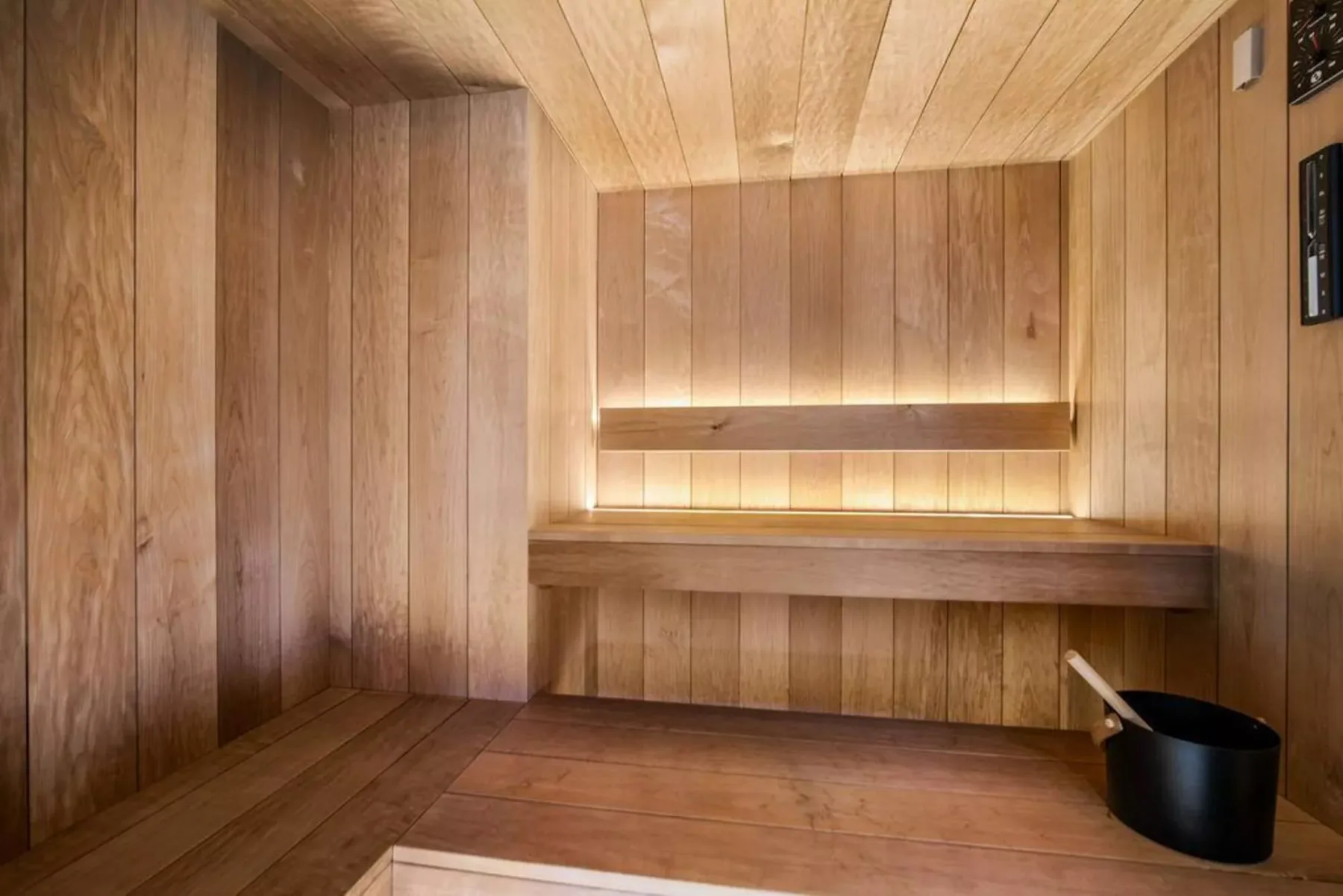 Una sauna revestida de madera con bancos escalonados y un cubo negro.