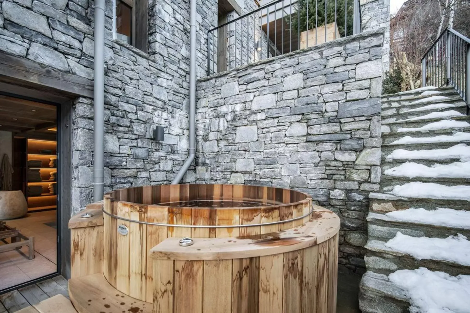 Una bañera de hidromasaje de madera está junto a muros de piedra y escalones cubiertos de nieve.