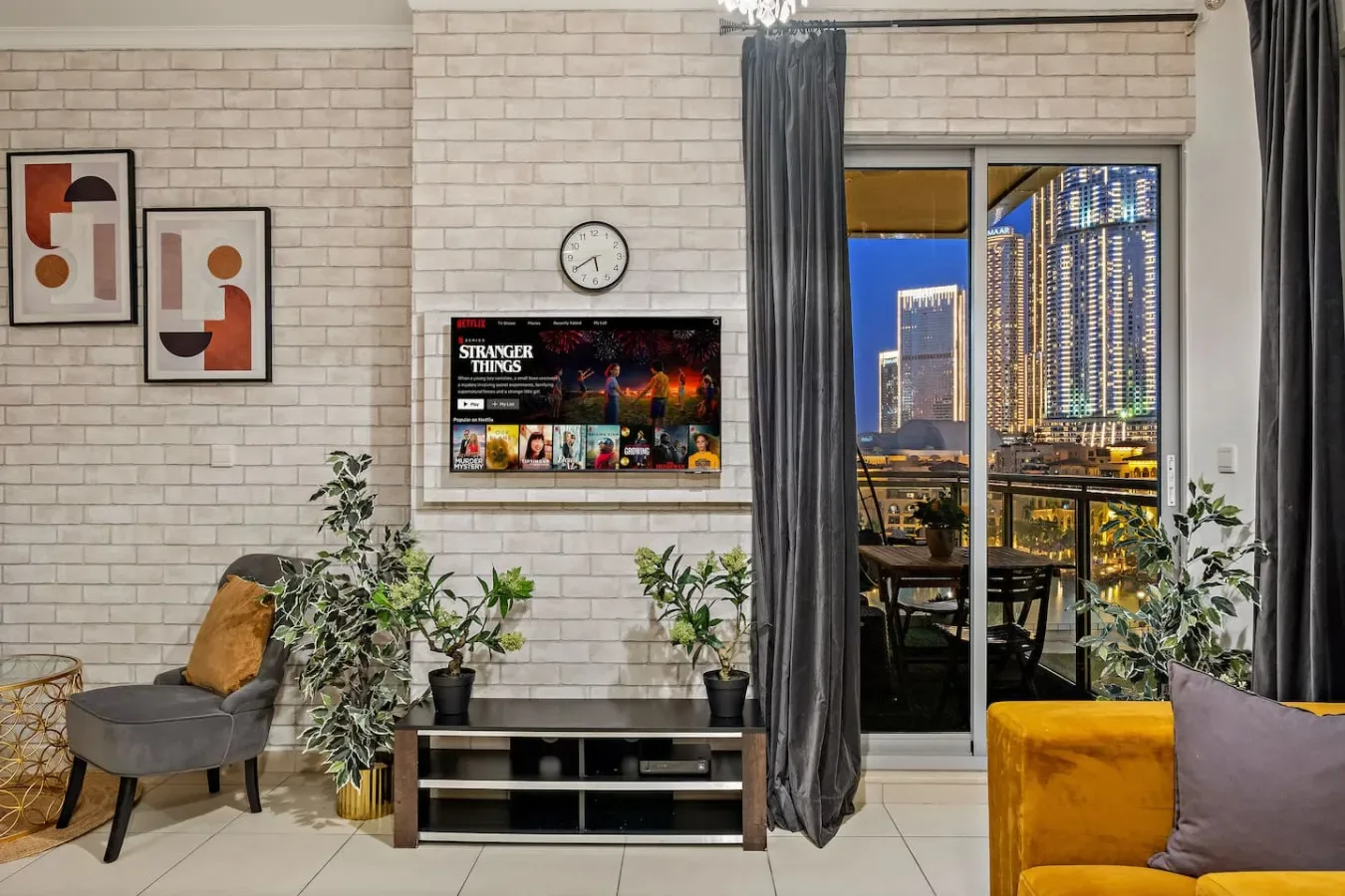 Una sala de estar con televisor, asientos y una gran ventana con vistas a las luces de la ciudad.