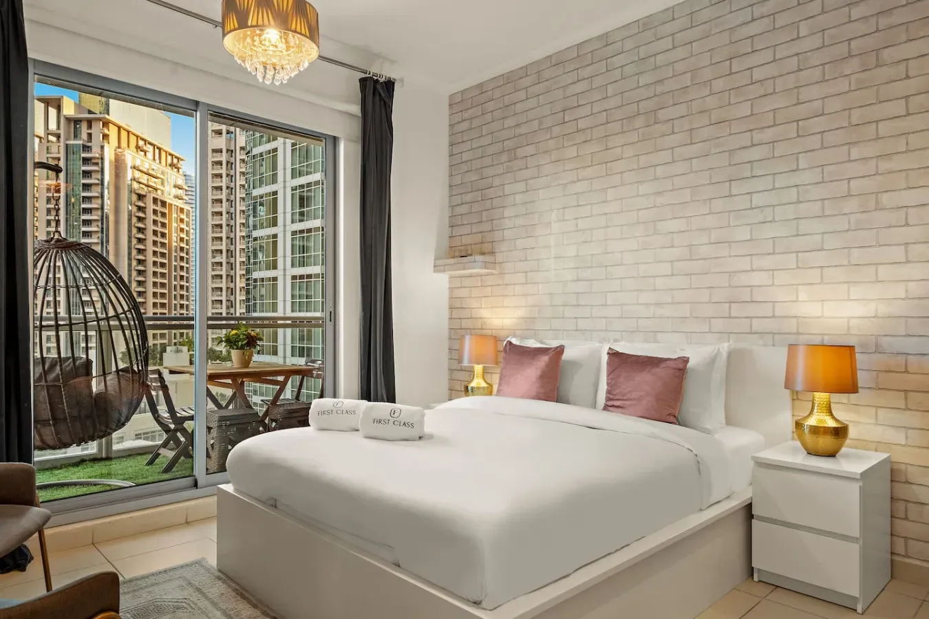Dormitorio con una cama grande, una pared de ladrillo claro y ventanas con vistas a edificios cercanos.