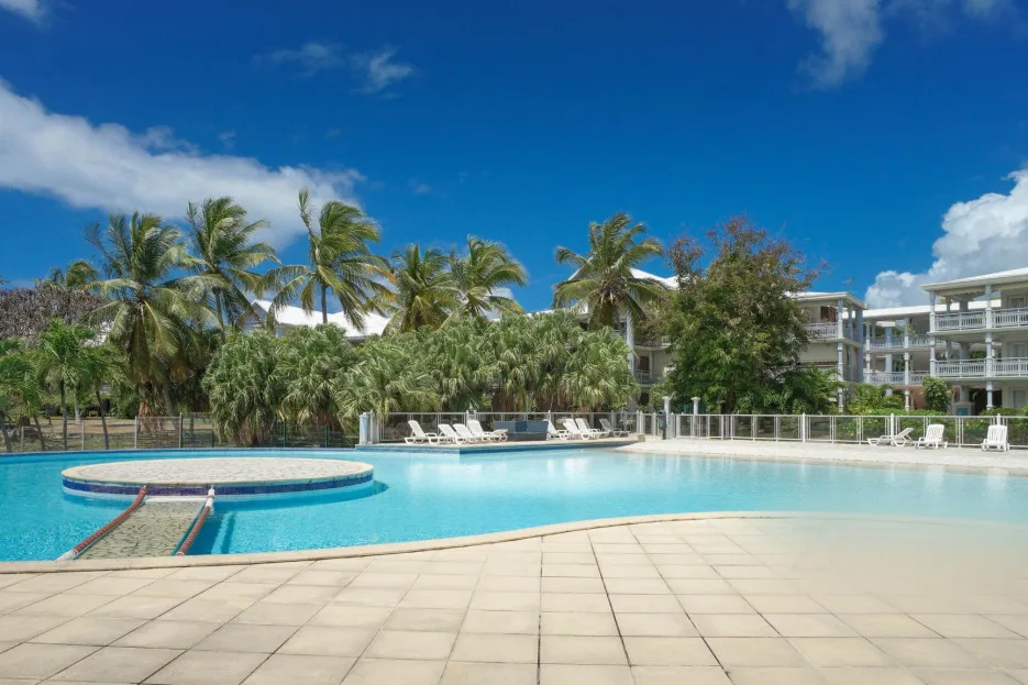 Una piscina de complejo turístico con una pequeña isla circular, palmeras y edificios al fondo.