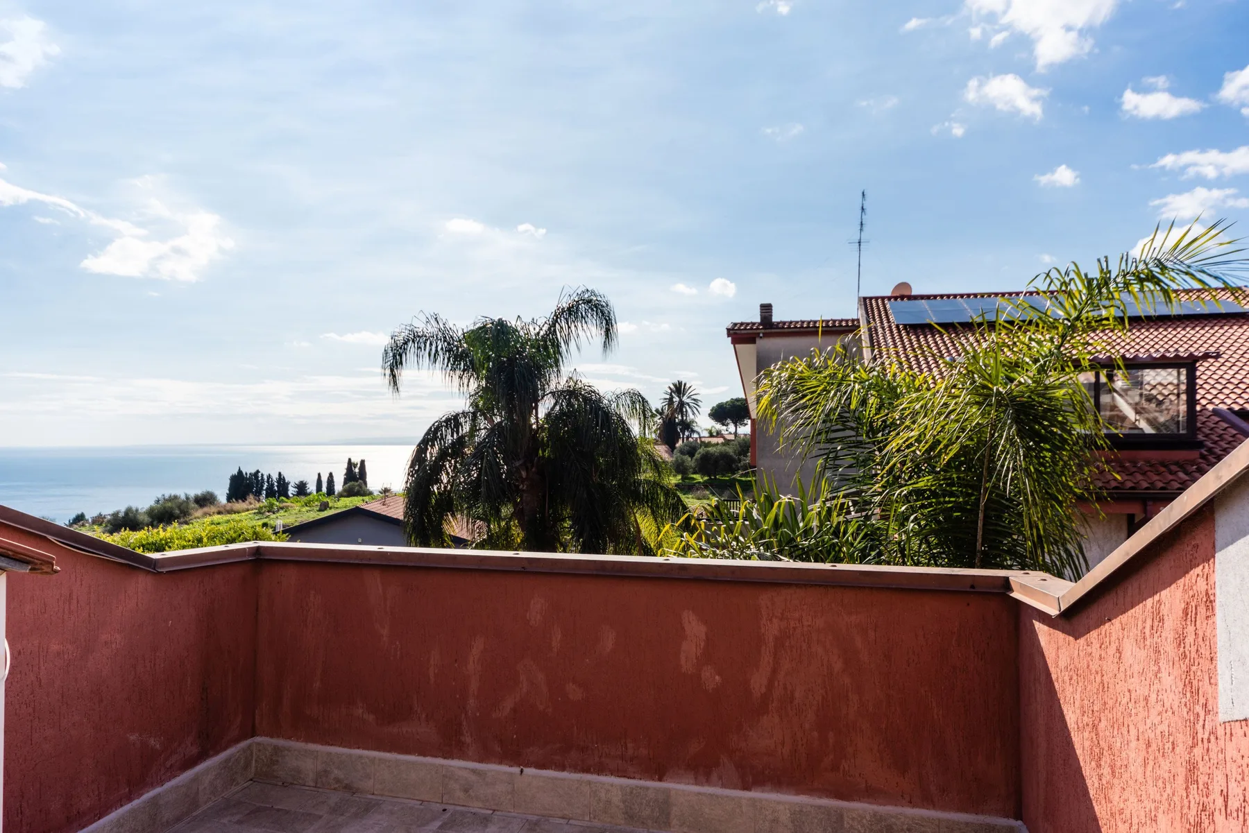 Una terraza da a casas cercanas, palmeras y una franja de mar a lo lejos.
