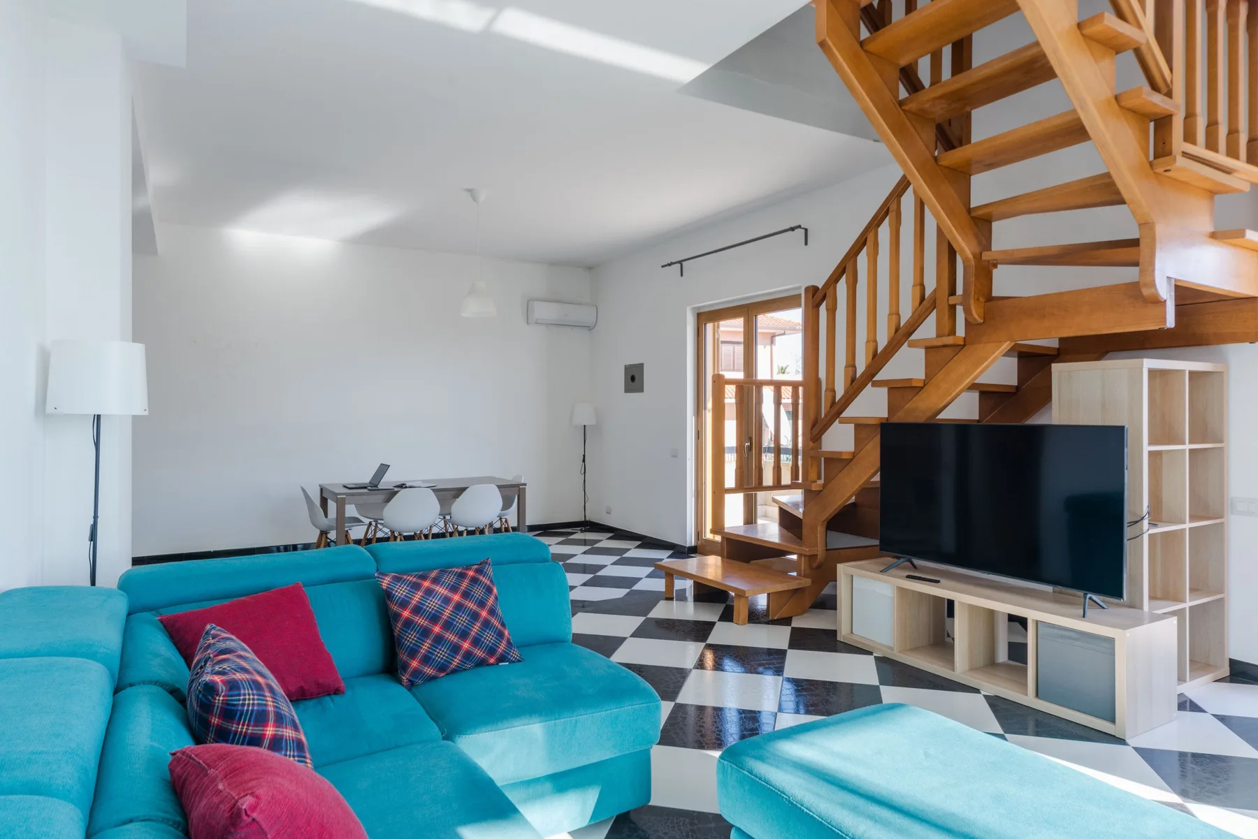 Una sala luminosa con asientos turquesa, un televisor y una escalera de madera.