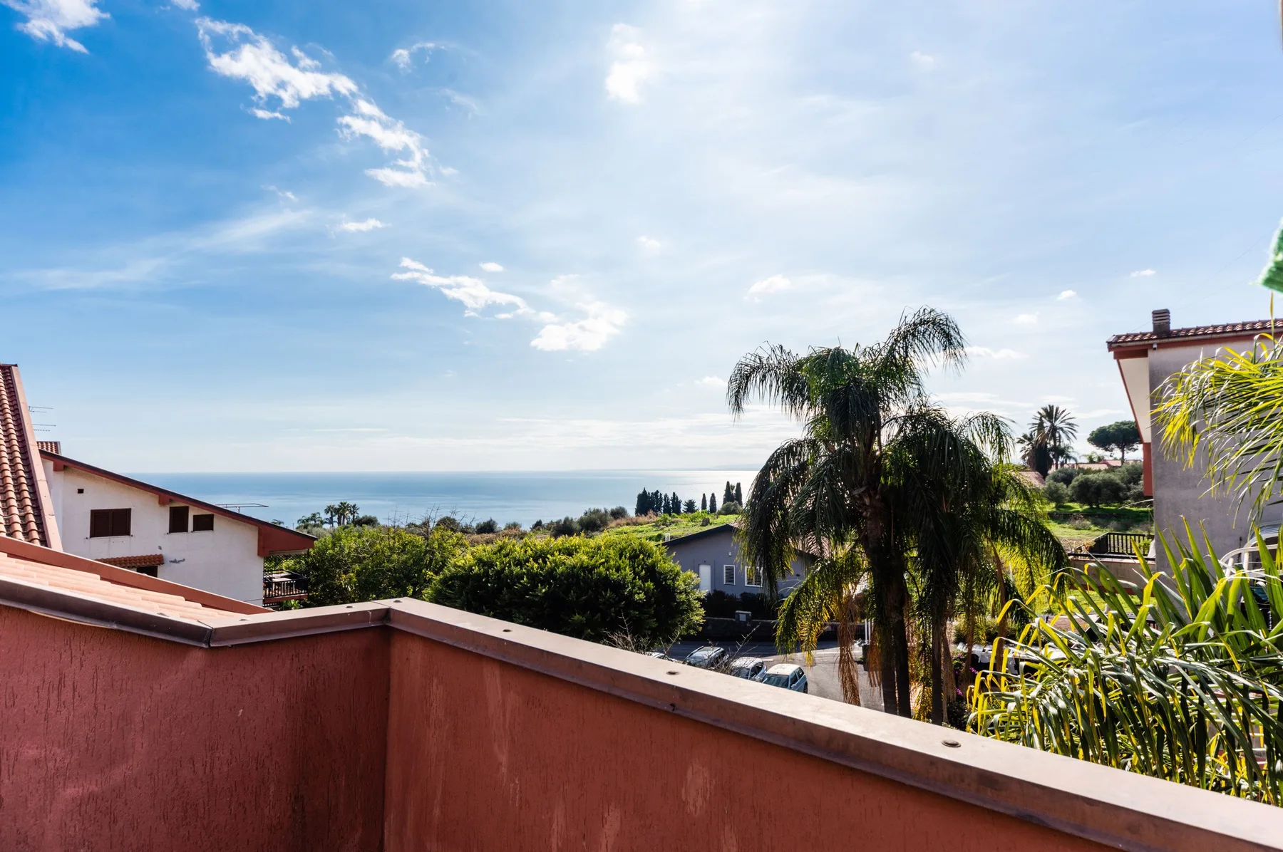Un balcón da a casas cercanas, palmeras y el mar bajo un cielo despejado.