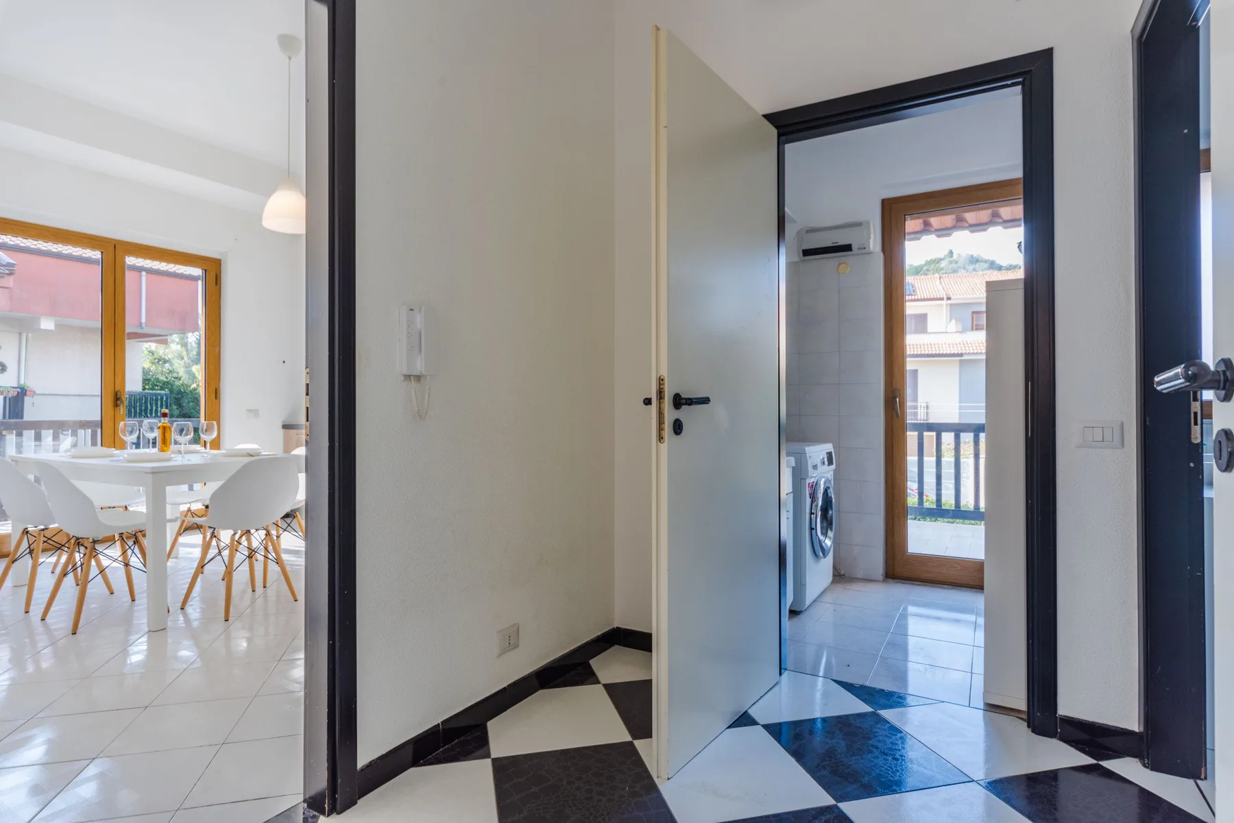 Una puerta abierta da a una habitación con suelo de baldosas, lavadora y puerta al balcón.