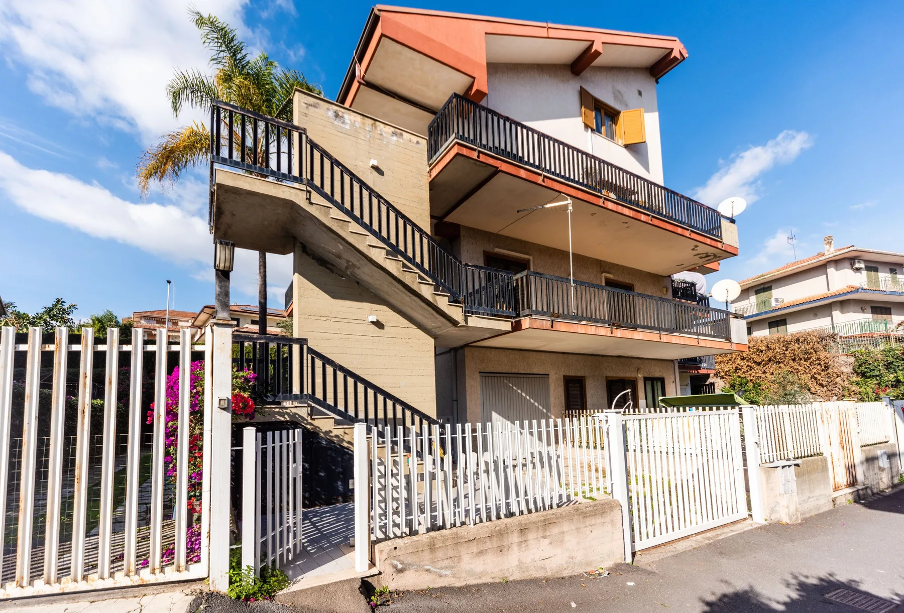 Una casa de varios pisos con balcones y una escalera exterior detrás de una cerca blanca.