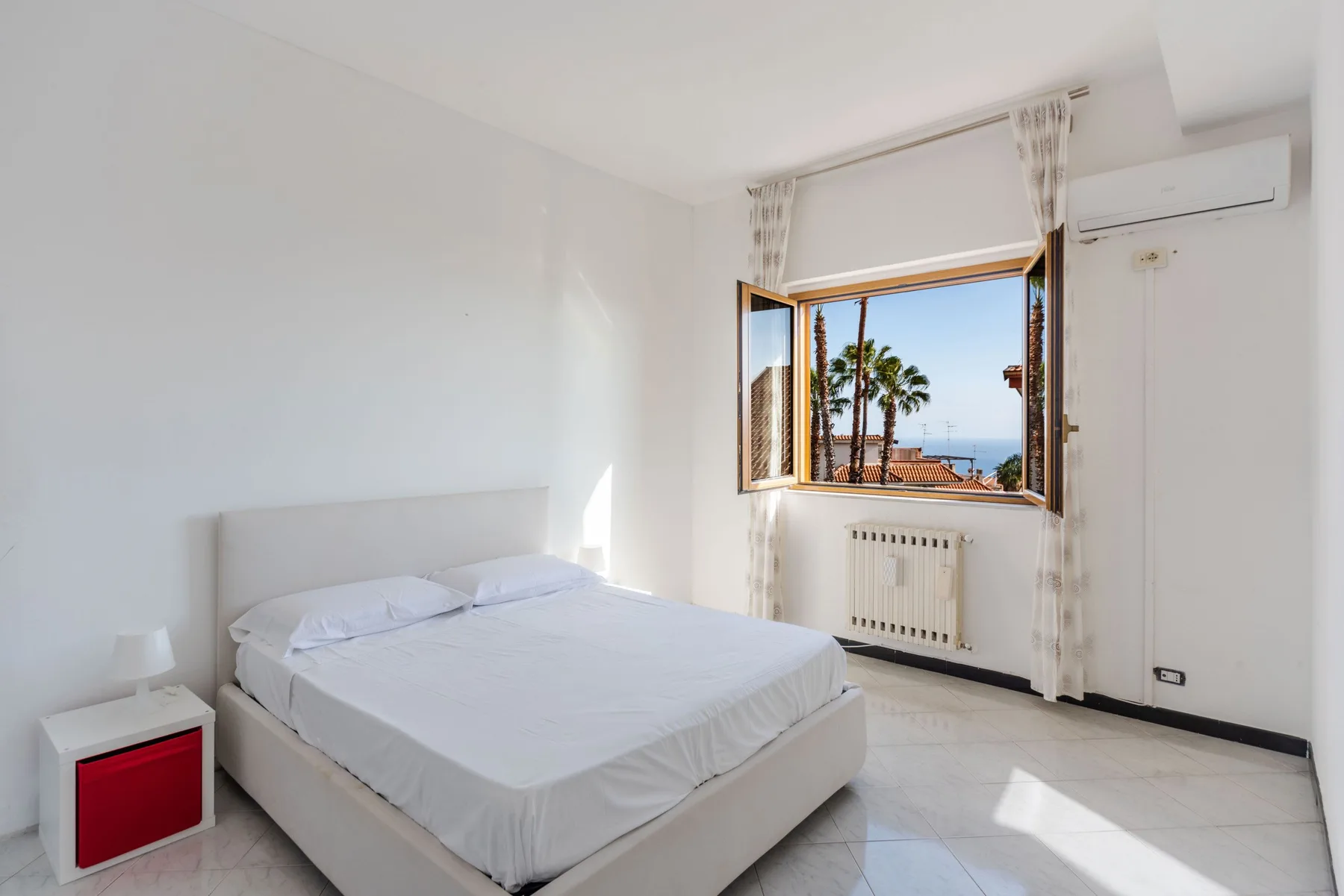 Un dormitorio luminoso con una cama y una ventana abierta con palmeras a la vista.