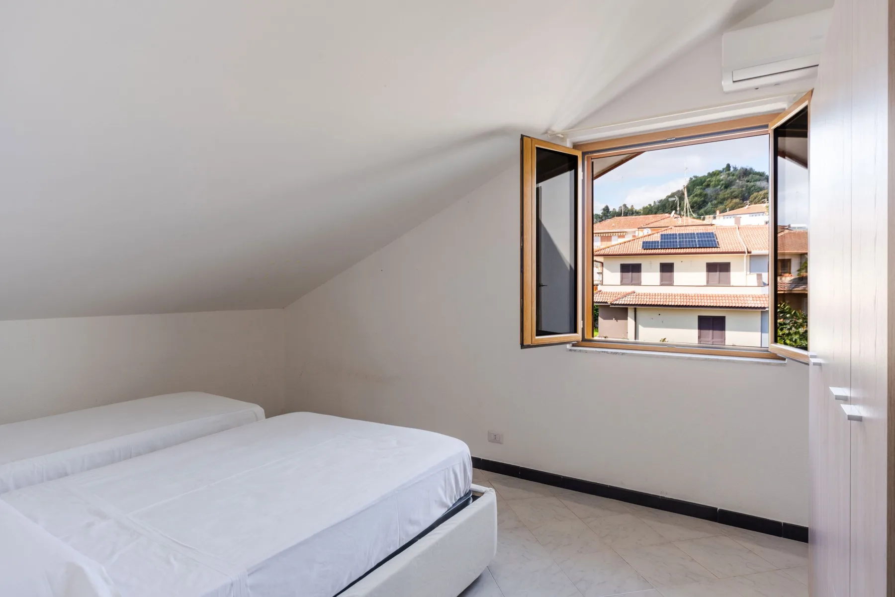 Un dormitorio luminoso con dos camas y una ventana abierta con vistas a edificios cercanos.