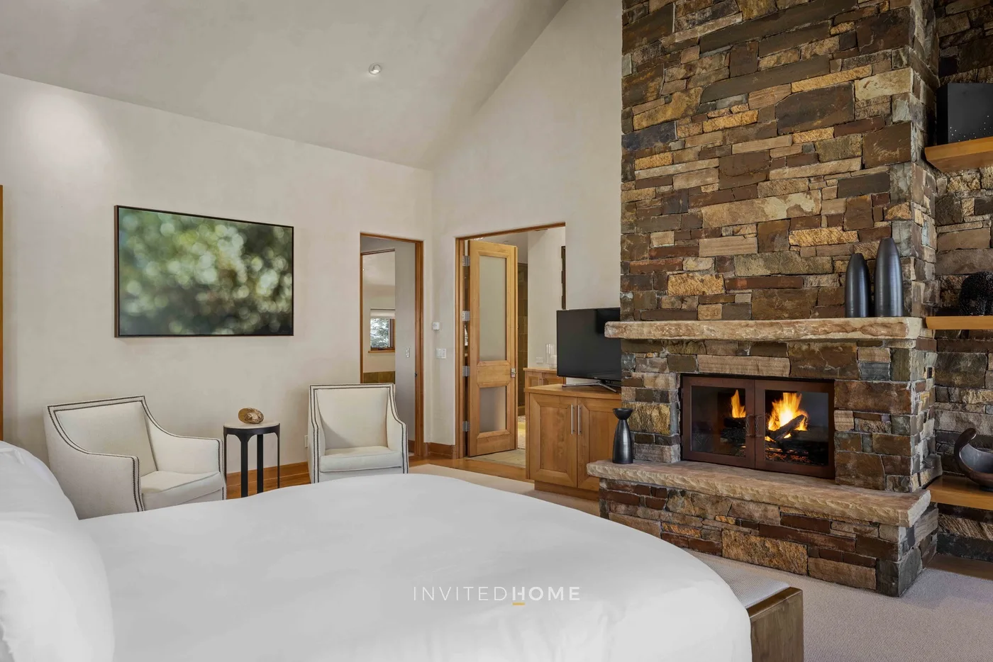 A bedroom with a large bed, two chairs, and a stone fireplace.