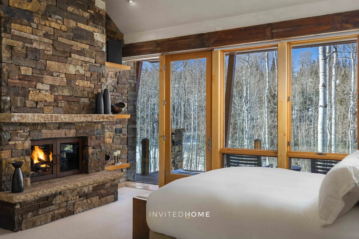 A bedroom with a bed beside a stone fireplace and large windows overlooking trees.