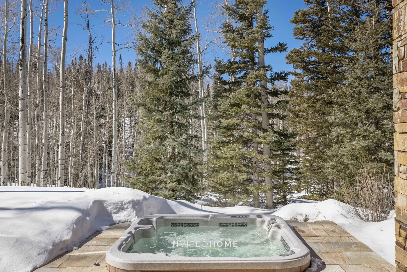 An outdoor hot tub sits on a stone patio beside snowbanks and trees.