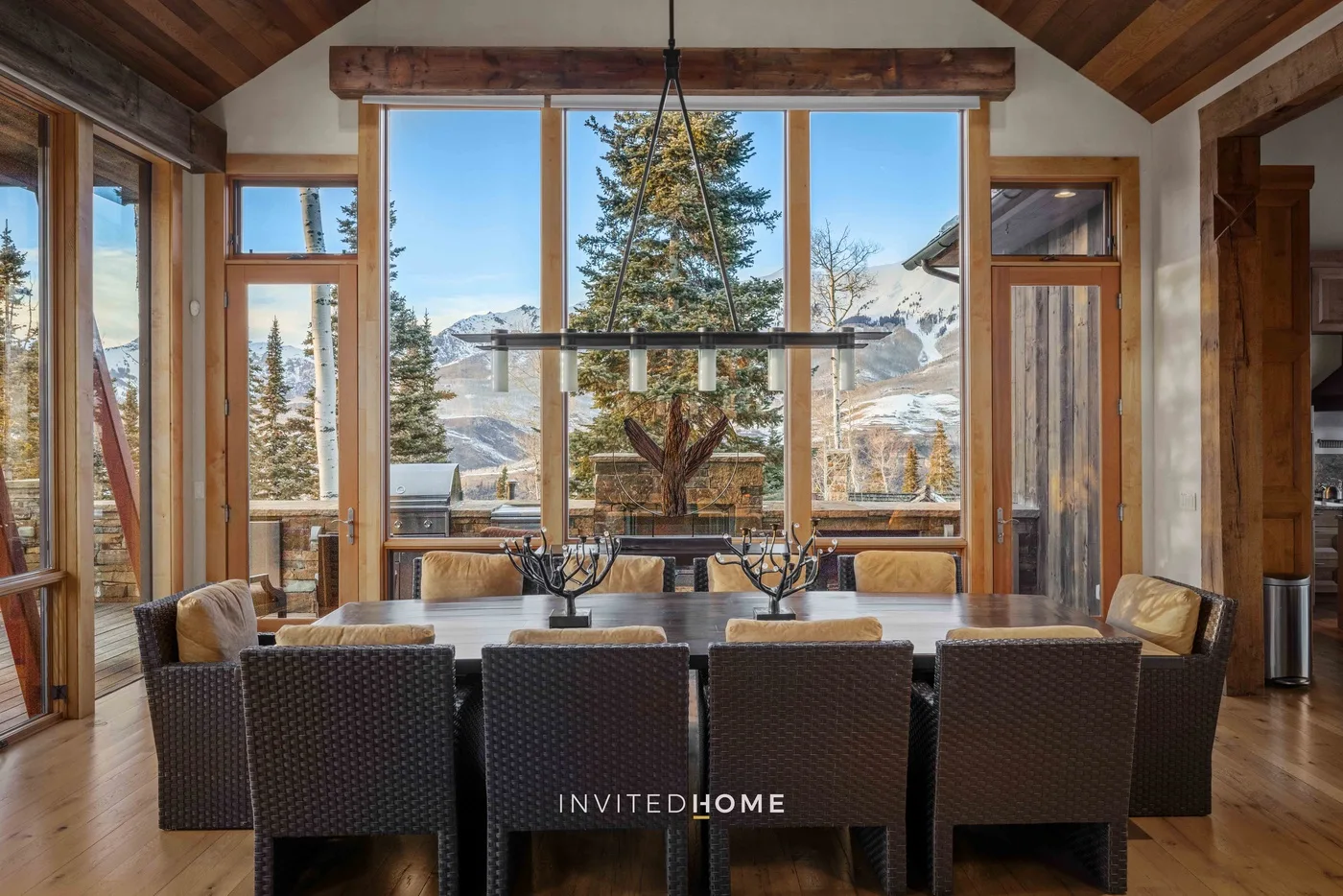 A dining area with a long table and dark woven seating faces large windows overlooking trees and mountains.