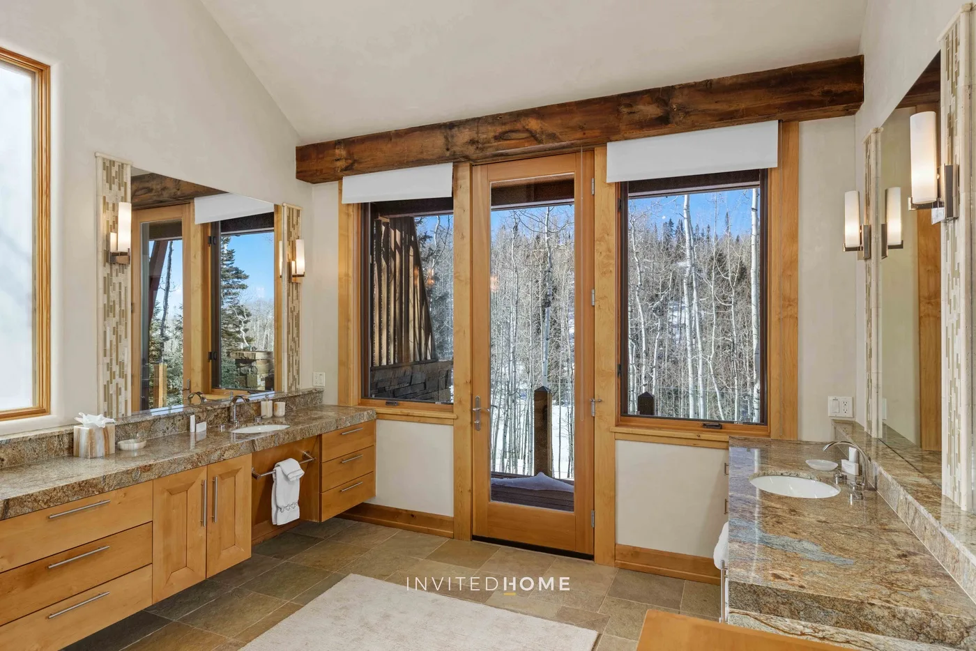A bathroom with a long vanity, sinks, and large windows overlooking trees.