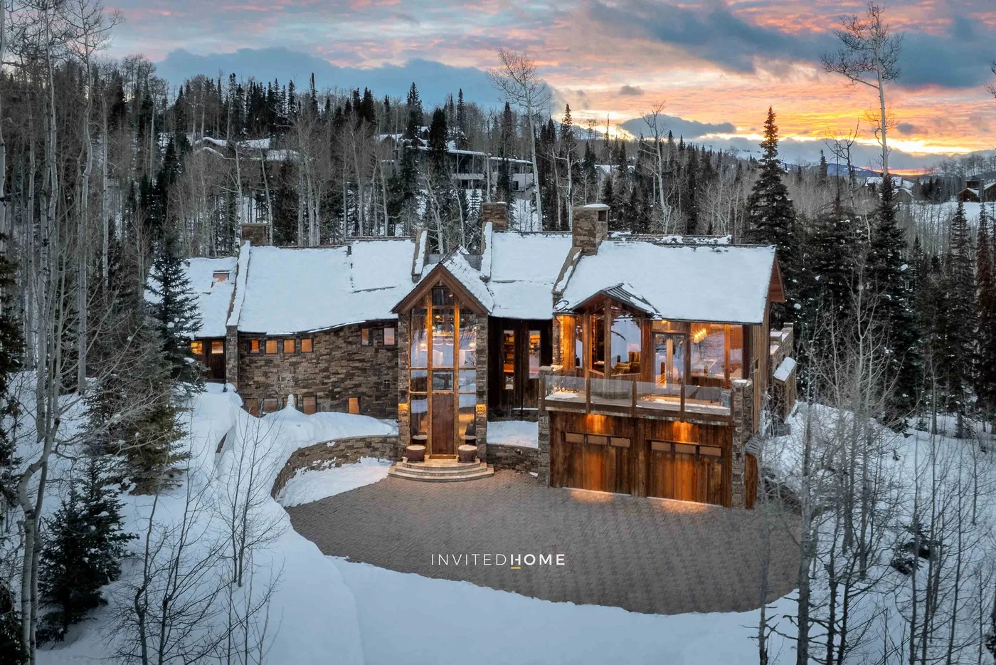 A snow-covered house sits among trees beneath a colorful sunset sky.