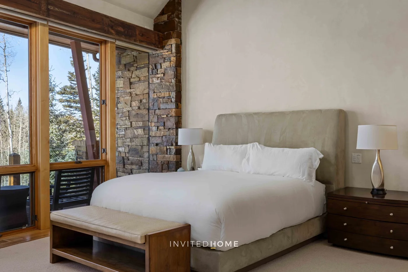 A bedroom with a neatly made bed, bedside lamps, and large windows.