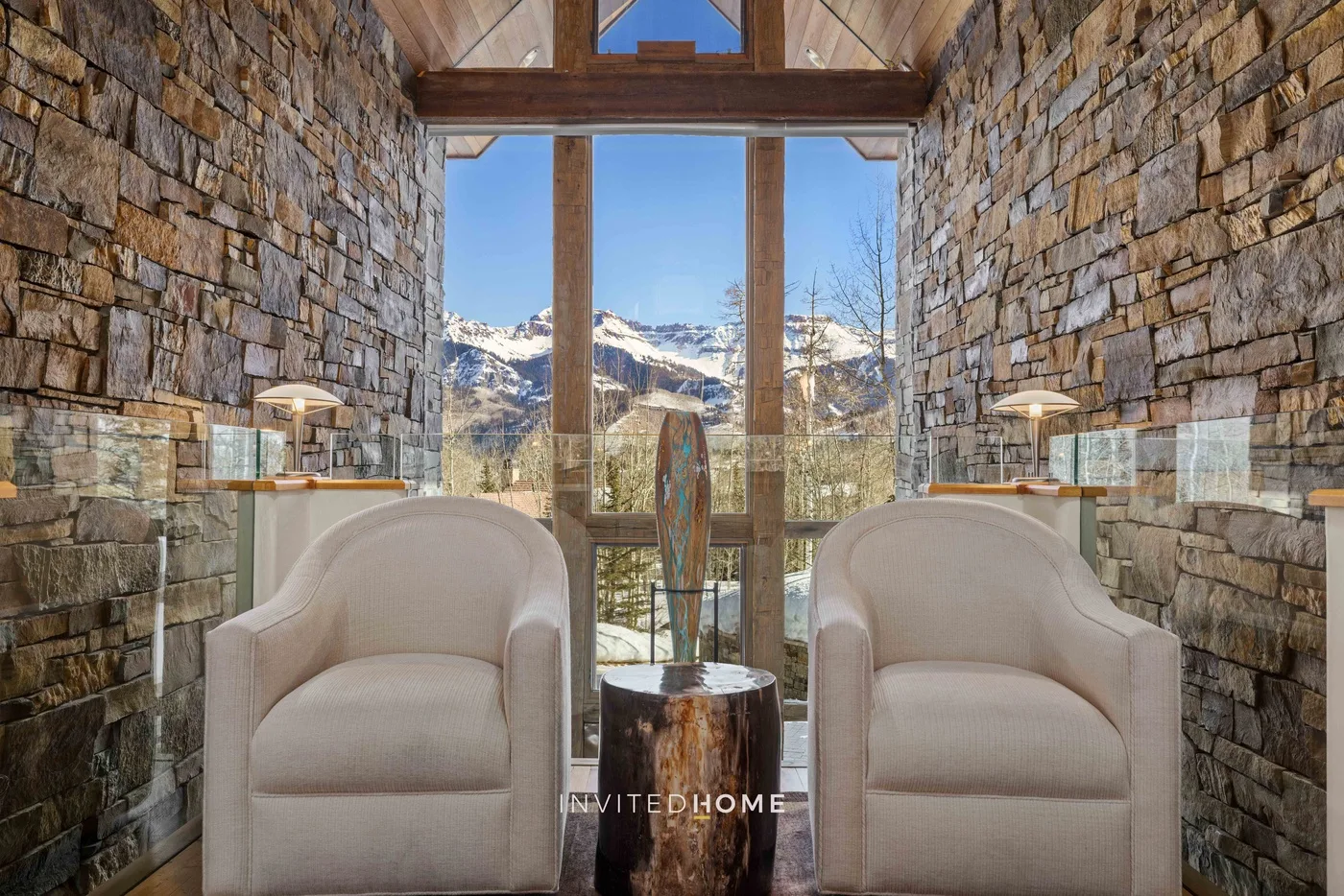 A stone-walled sitting area with two upholstered chairs and a large window overlooking mountains.