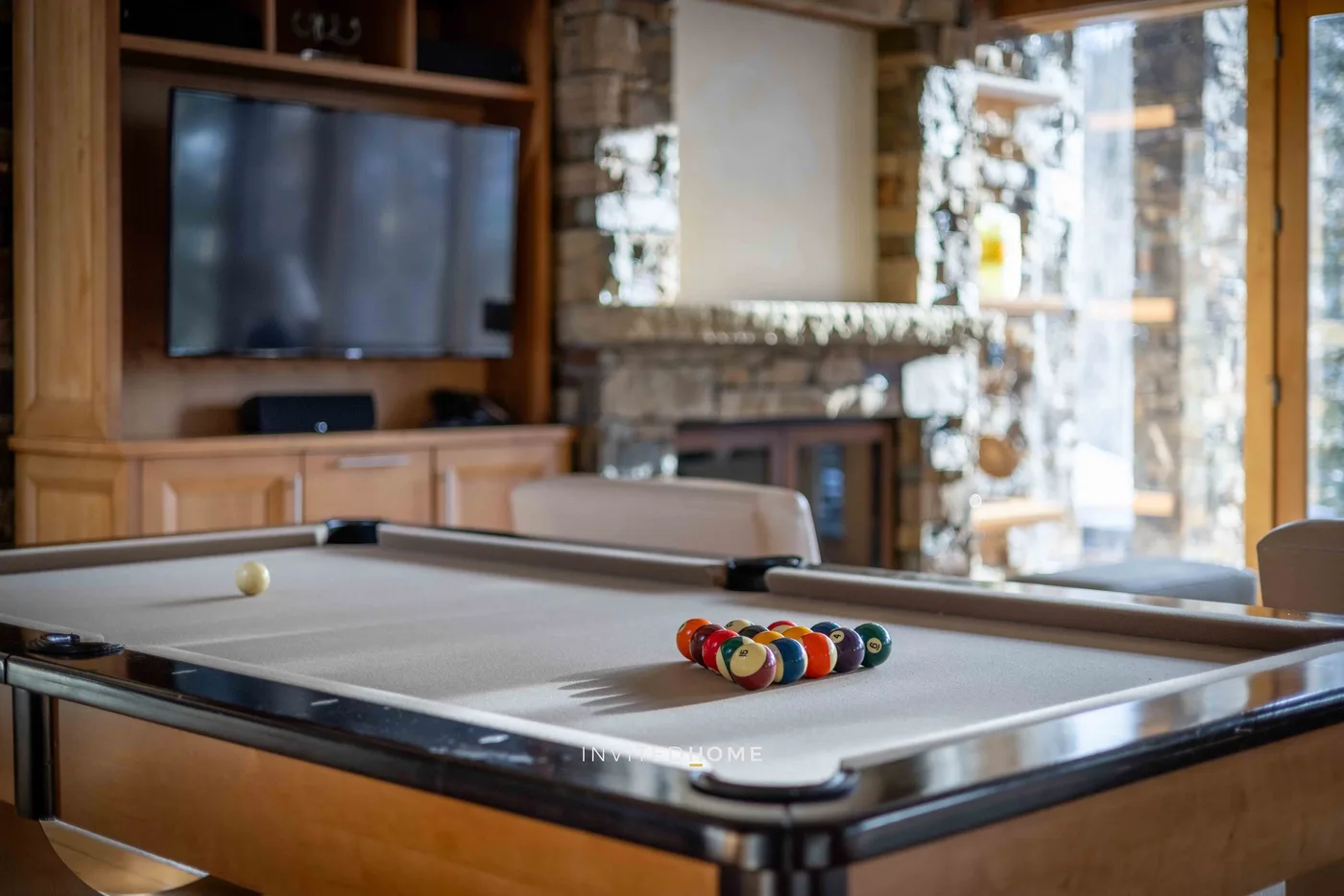 A billiards table stands in a living room with a television and stone fireplace.