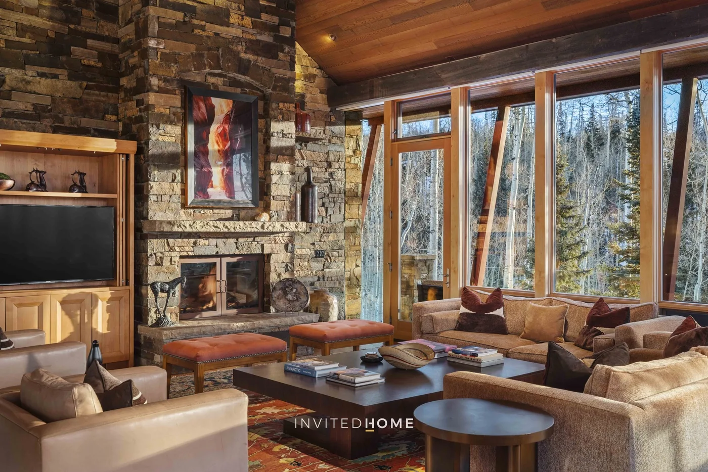 A living room with a stone fireplace, seating, and large windows overlooking trees.