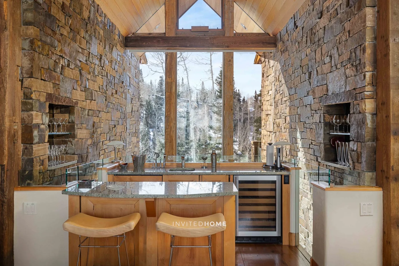 A stone-walled kitchen with a central island and tall windows overlooking trees.