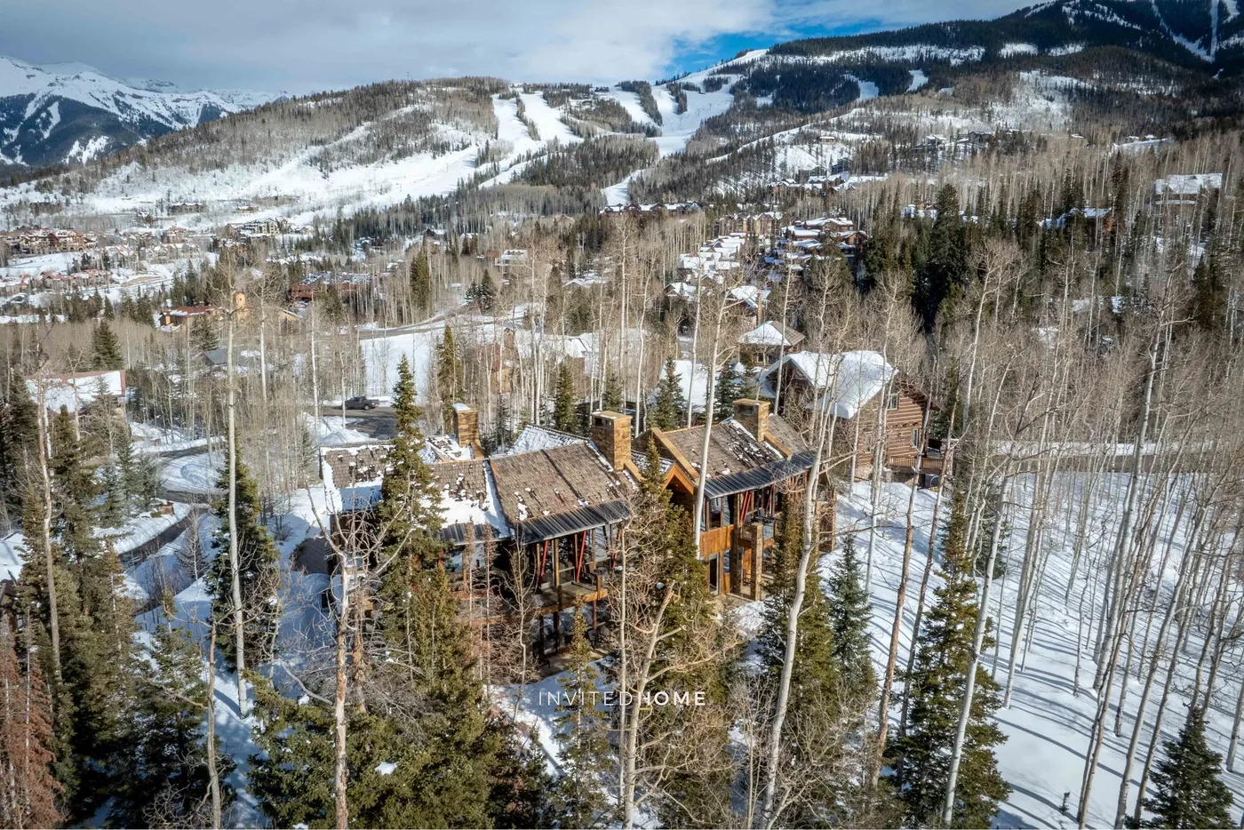 Aerial view of cabins among snowy trees, with ski slopes across the mountain in the background.