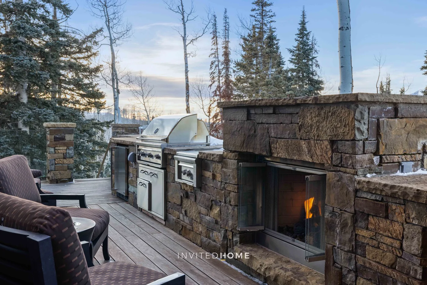 A stone outdoor kitchen and fireplace sit beside deck seating among evergreen trees.