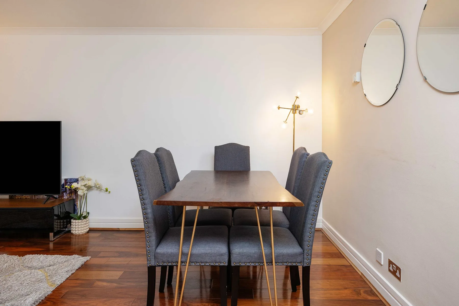 A dining table with upholstered seating sits beside a television in a bright room.