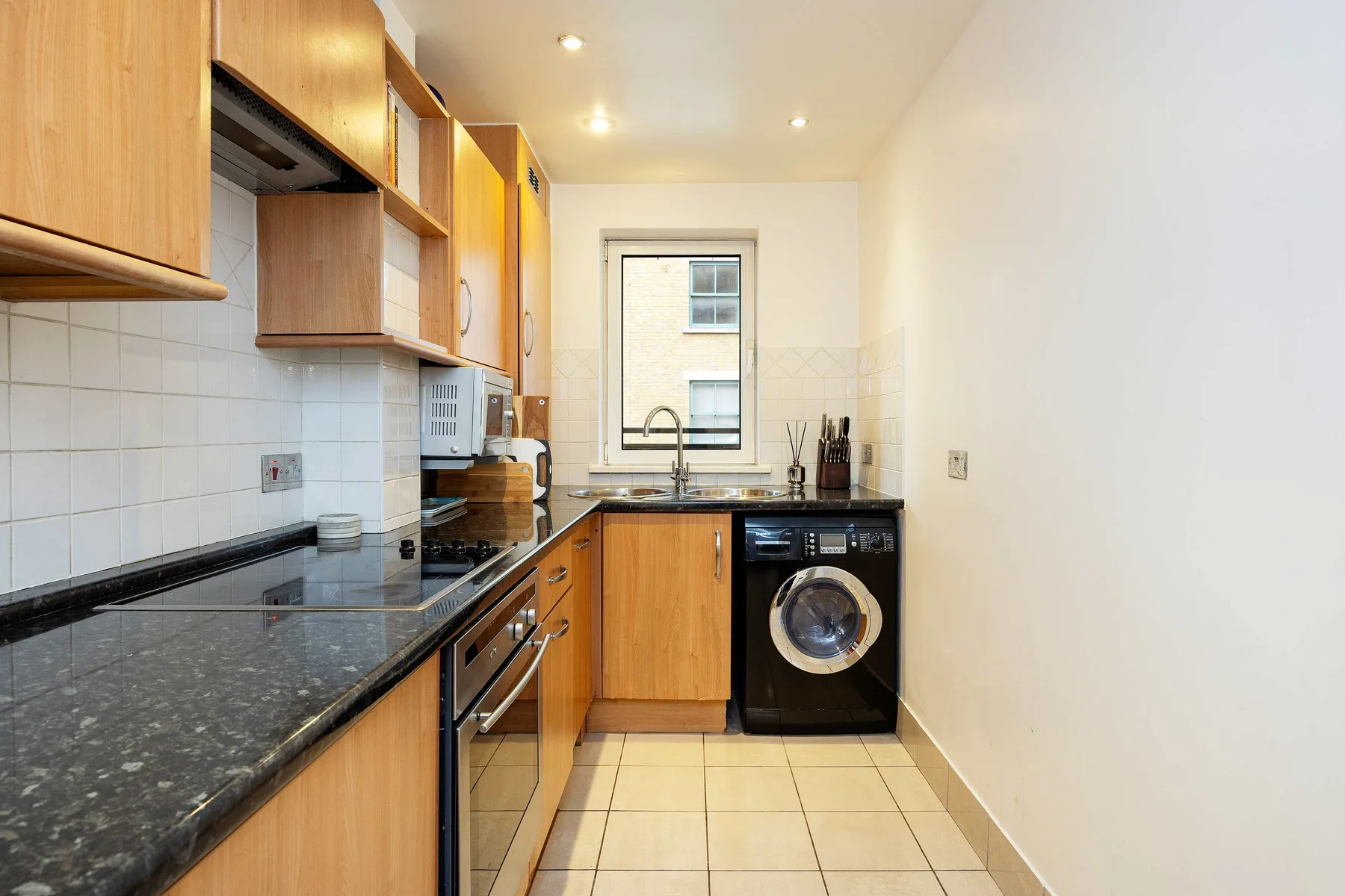 A narrow kitchen with wooden cabinets, dark countertops, and a washing machine.