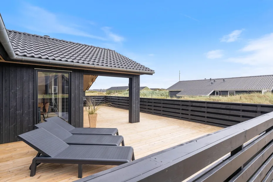 A wooden deck with reclining lounge chairs overlooks neighboring rooftops under a blue sky.