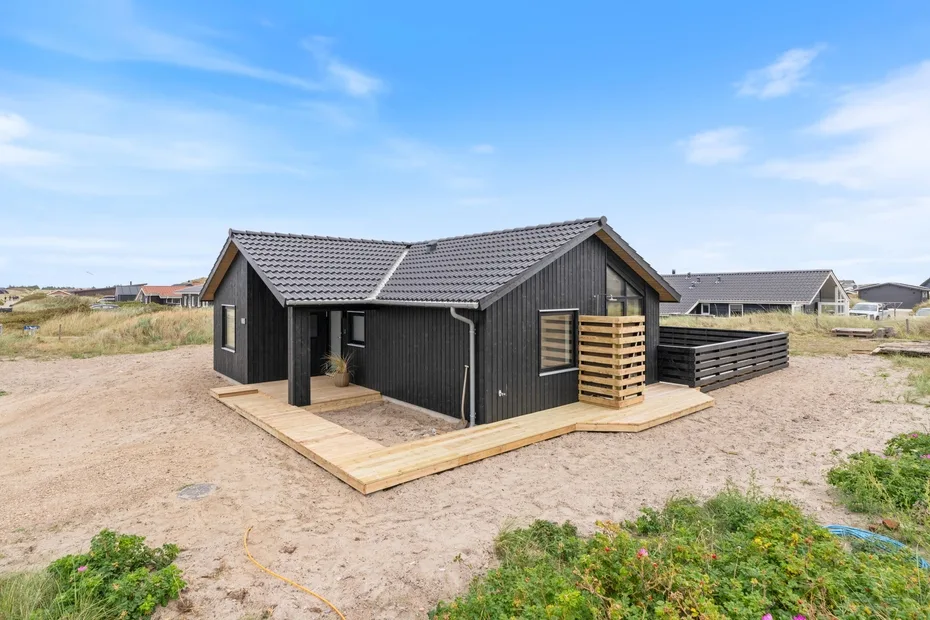 A dark beachside house with a wooden deck stands among sandy dunes.