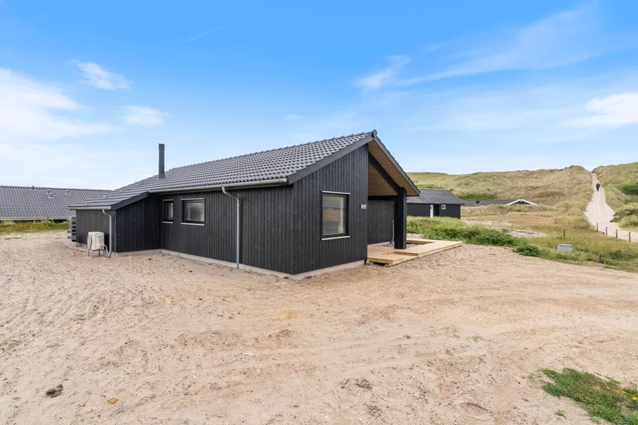 A dark-sided house stands on sandy ground beside grassy dunes.