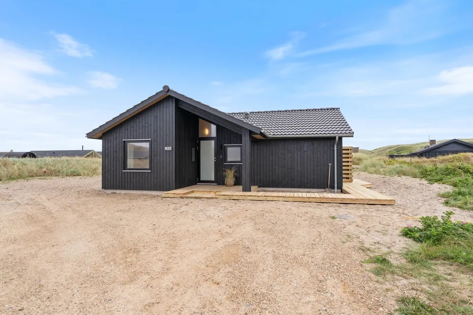 A dark, single-story house stands in an open sandy landscape.