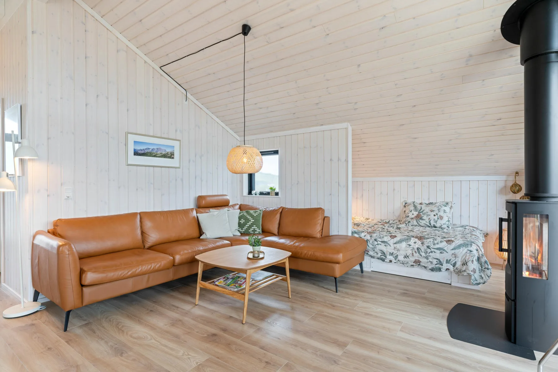 A bright living area has an L-shaped seating arrangement beside a bed and wood stove.