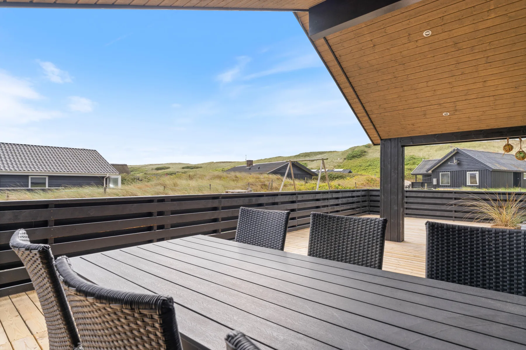 A covered outdoor dining area with a dark table and woven seating overlooks grassy dunes.