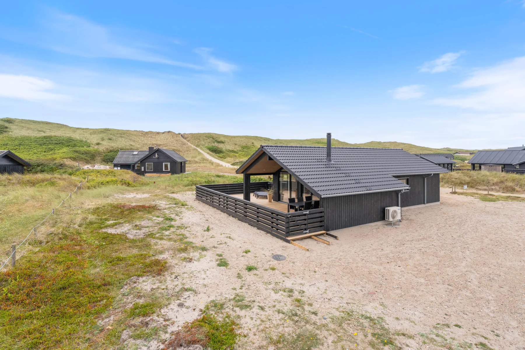 A dark house with a fenced deck stands among sandy paths and grassy dunes.