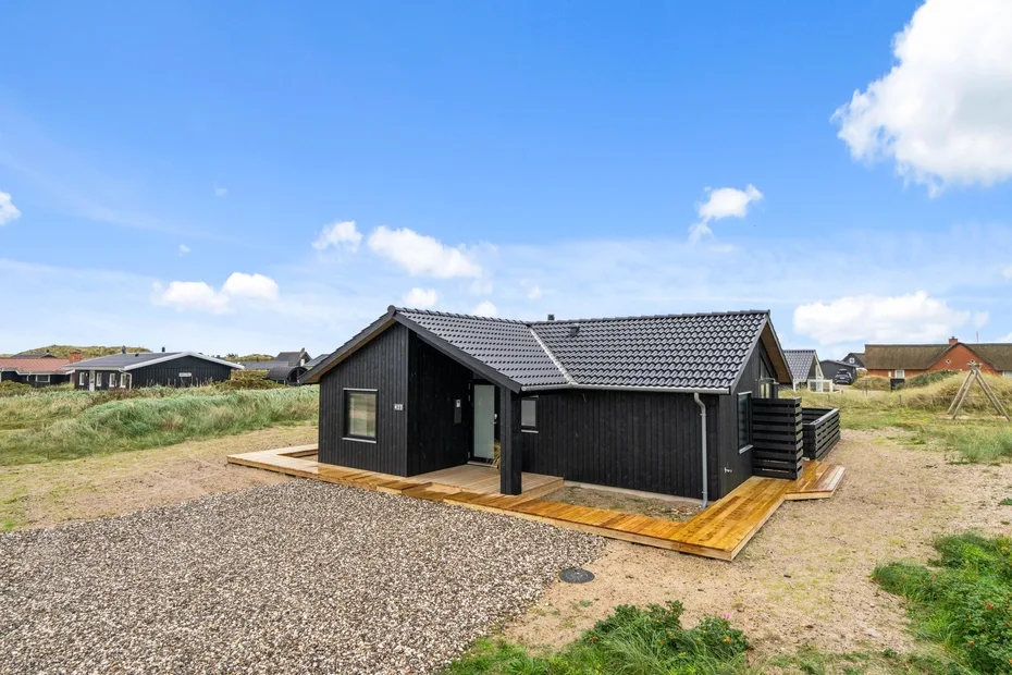 A dark-sided house stands on a wooden deck in an open grassy landscape.