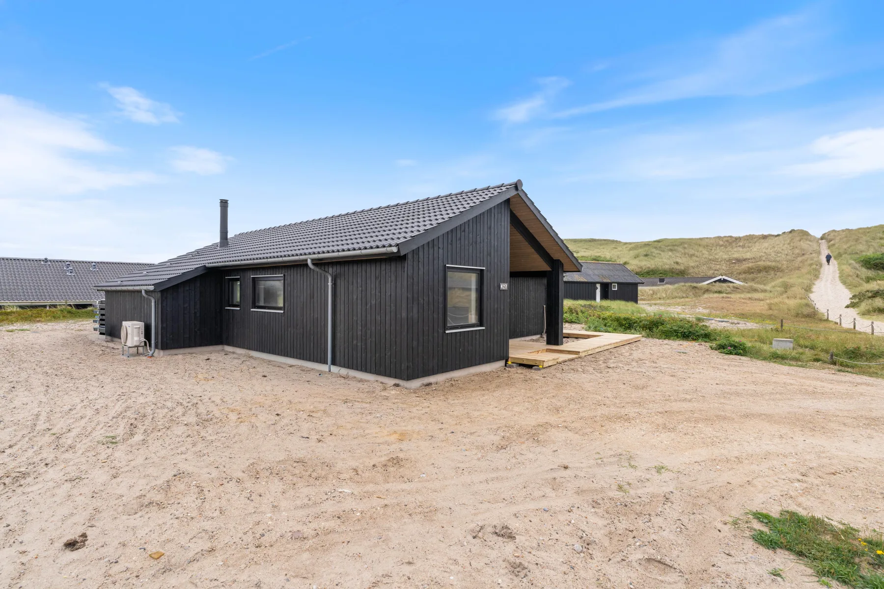A dark wooden house stands on a sandy lot beside grassy dunes.