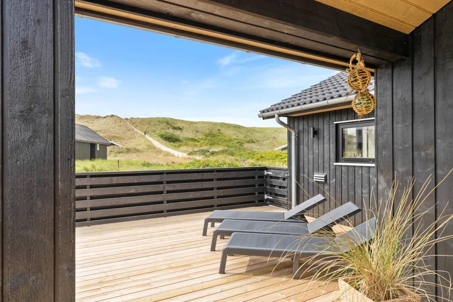 A wooden deck with lounge seating overlooks grassy dunes beside a dark-sided building.