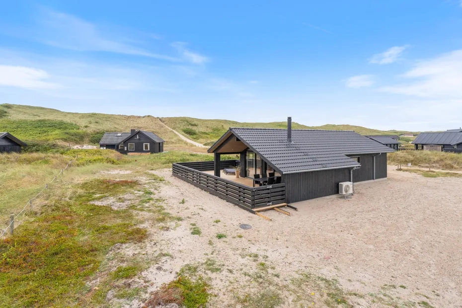 A dark cabin sits beside sandy ground and grassy dunes.