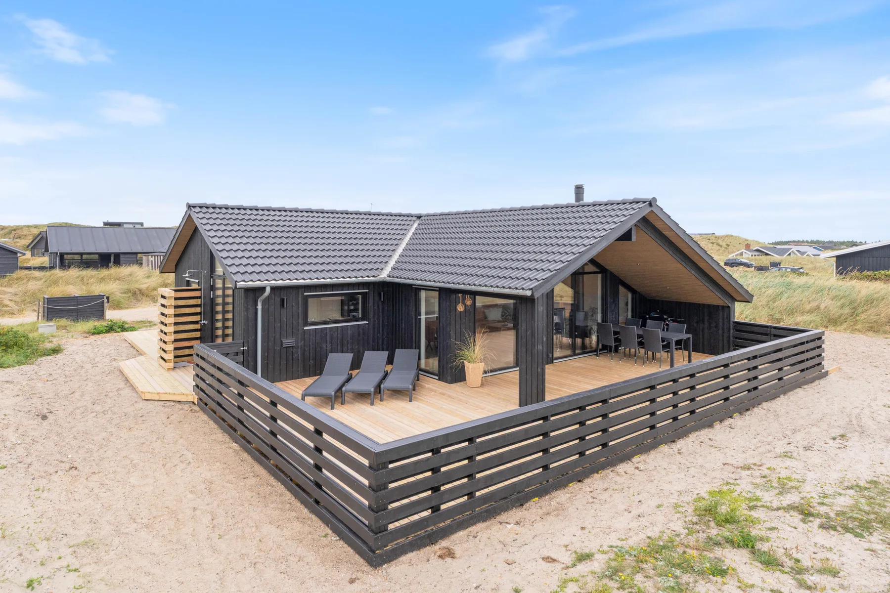 A dark-sided vacation house with a fenced wooden deck sits among sandy dunes.