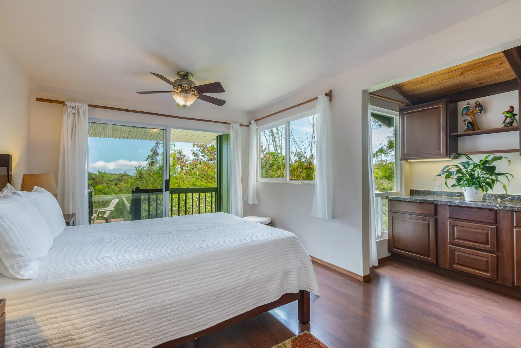 A bright bedroom with a large bed, wide windows, and a wooden cabinet.