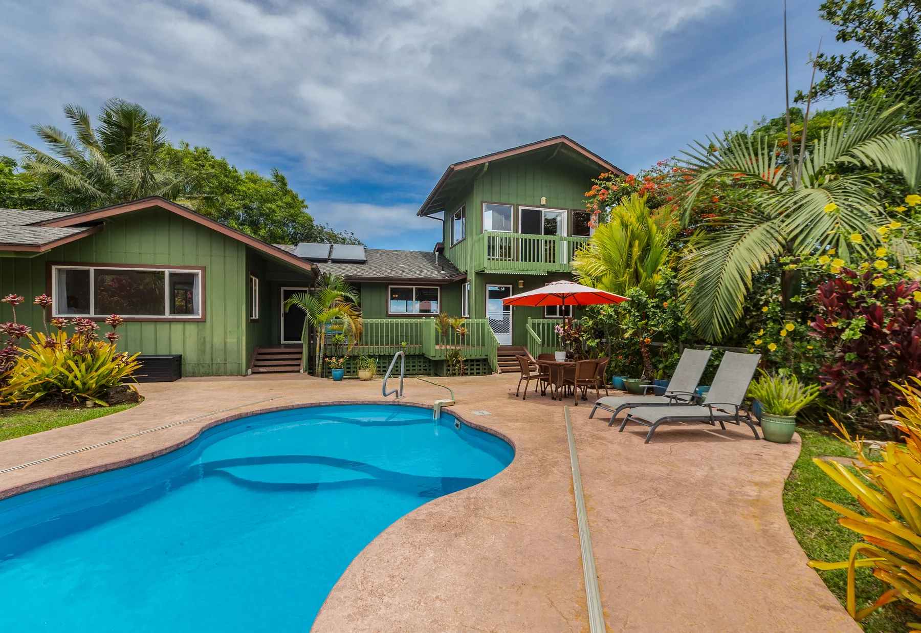 A swimming pool and patio sit in front of a green house amid tropical plants.