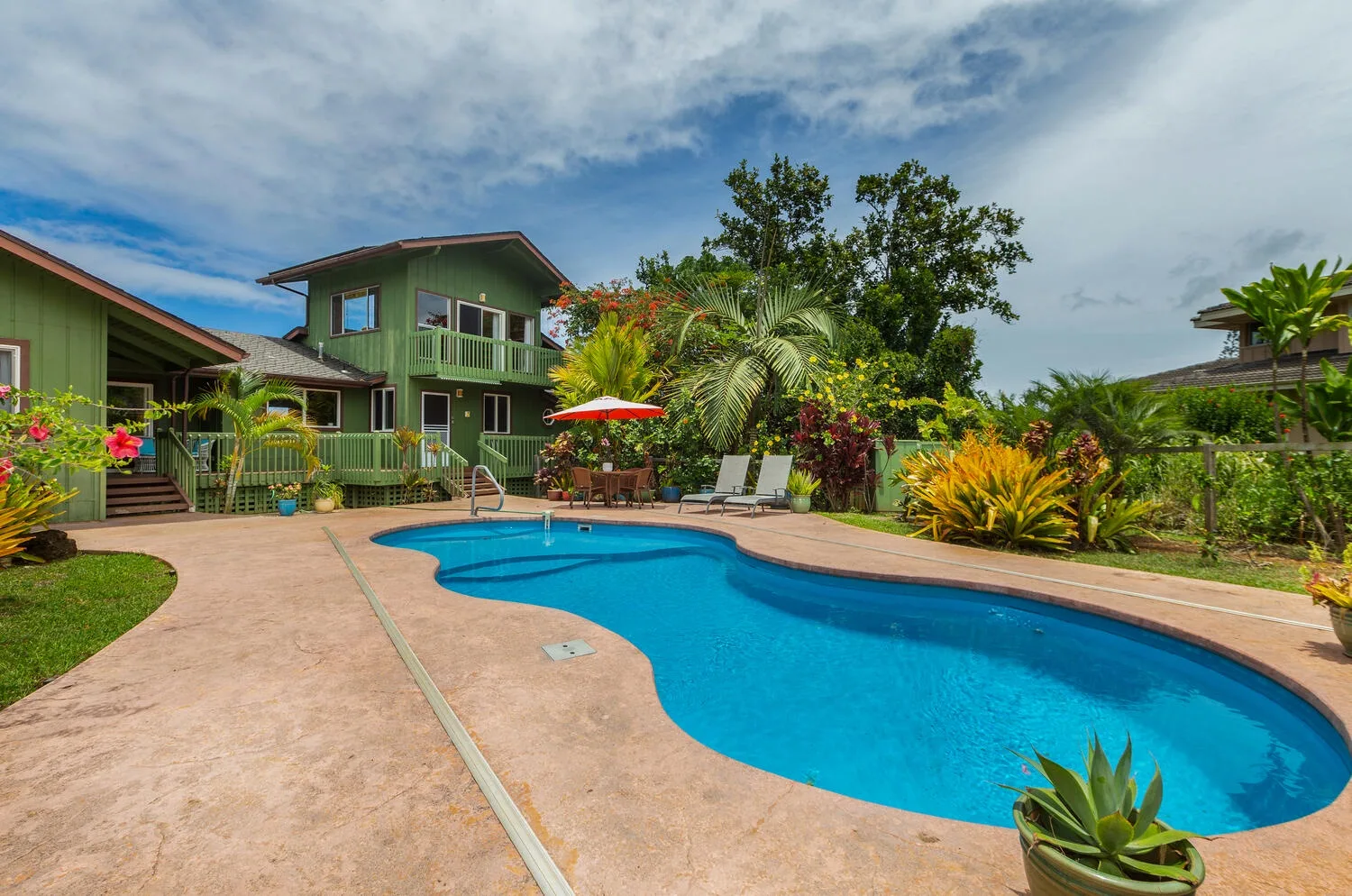 An outdoor swimming pool sits beside green buildings and tropical plants.