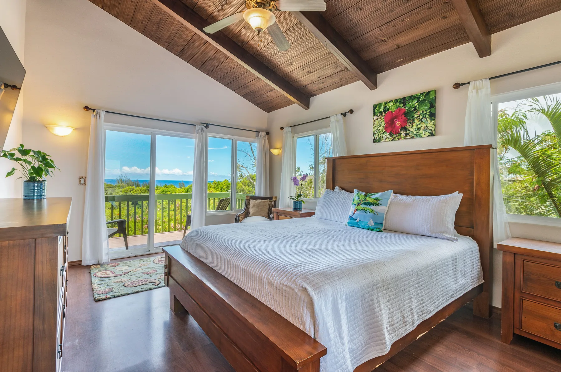 A bedroom with a large bed, wood-paneled ceiling, and windows overlooking greenery.