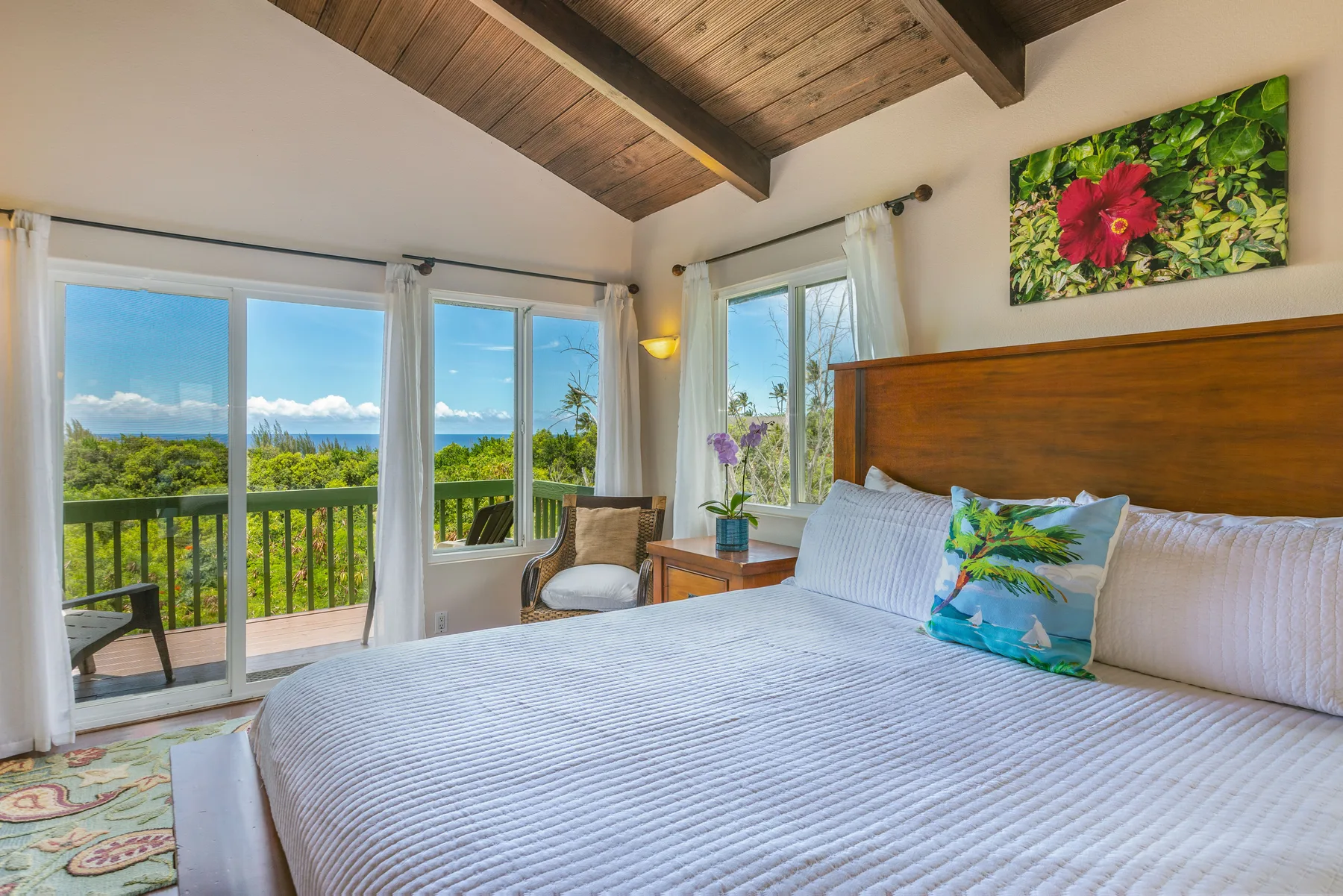 A bright bedroom with a large bed beside windows overlooking green trees and the ocean.