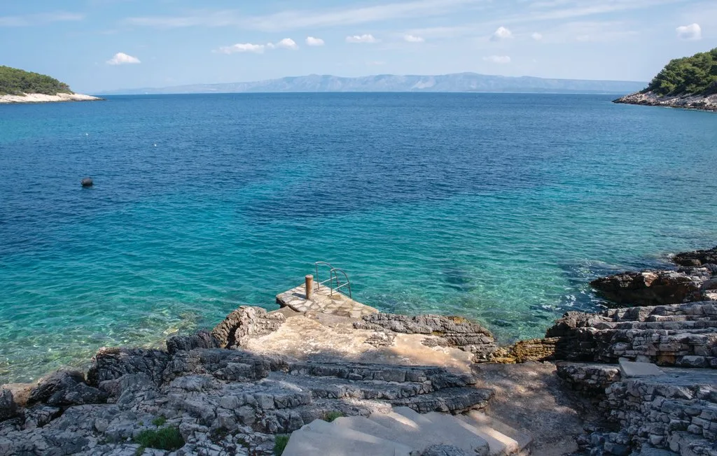 Una costa rocosa se abre hacia aguas azules y claras, con colinas lejanas en el horizonte.
