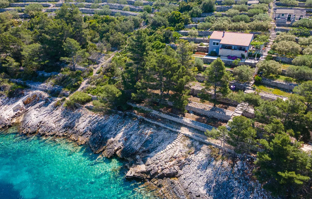 Vista aérea de una costa rocosa con agua turquesa clara y una casa entre árboles.