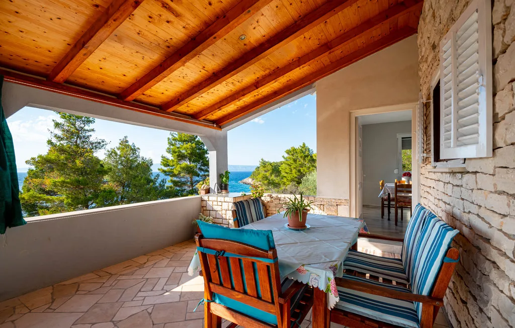 Comedor exterior cubierto con mesa y asientos, con vistas a árboles y al mar.