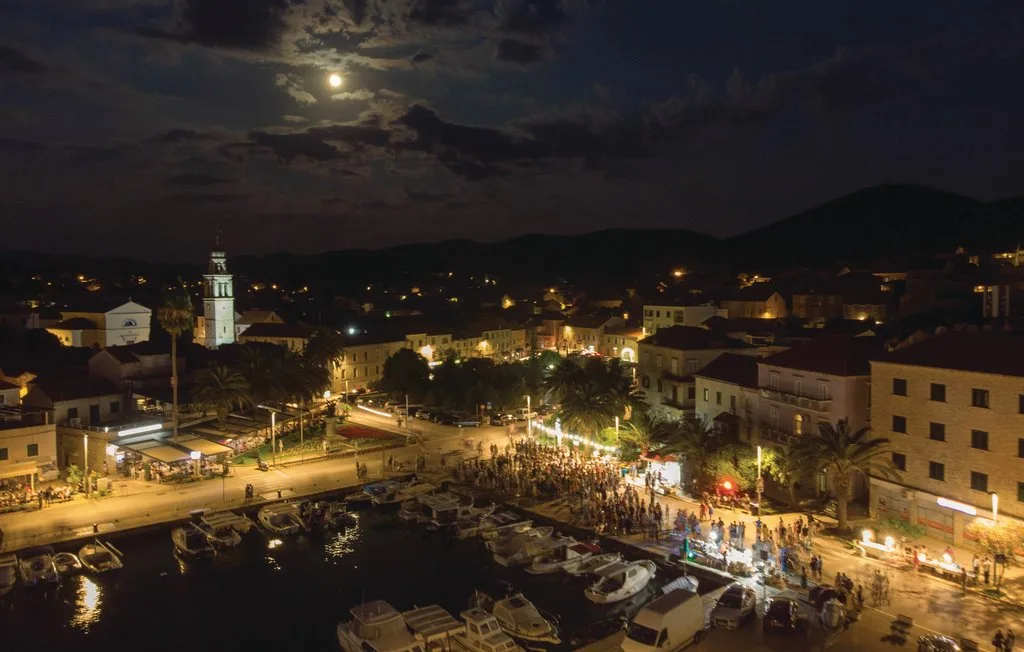 Una plaza junto al agua, iluminada por la luna y farolas, está junto a un puerto concurrido.