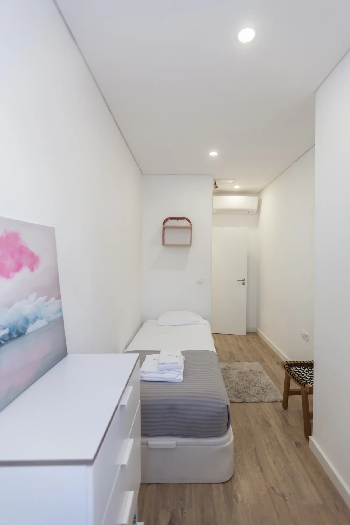 A compact bedroom with a single bed, white dresser, and a door at the far end.