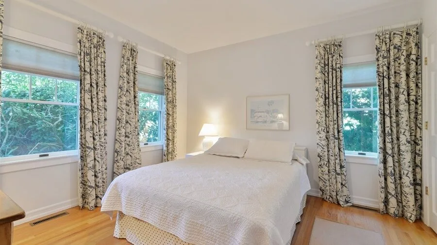 Un dormitorio con una cama bien tendida, cortinas estampadas y ventanas con vista a los árboles.